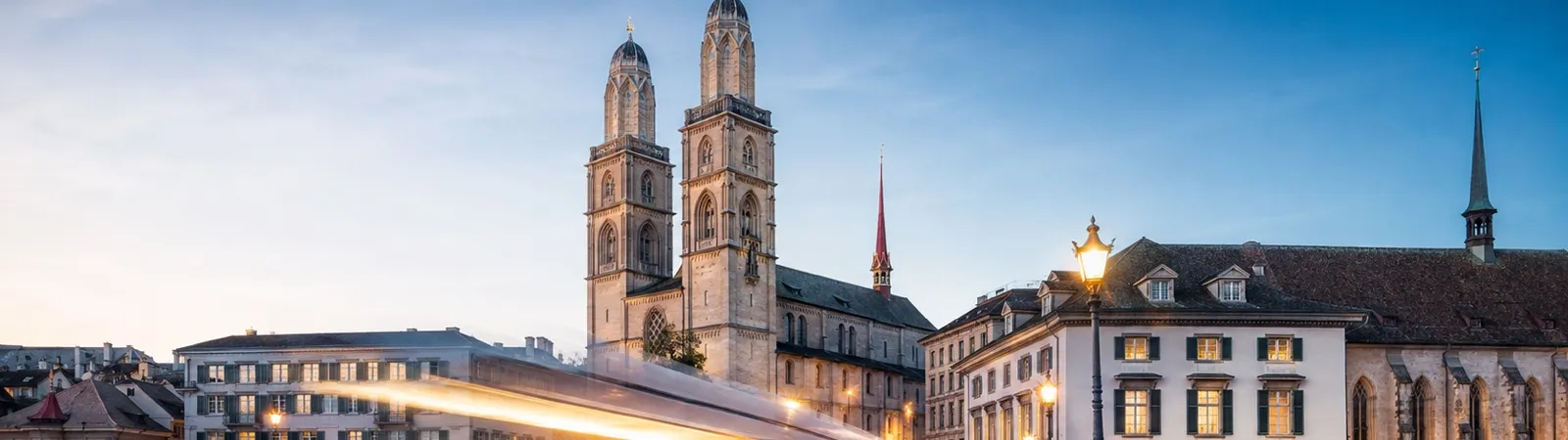 Zurich at dusk: the Grossmünster towers above the Limmat, with a tram passing as a streak of light.